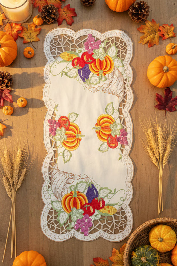 An autumn-themed embroidered table runner with scalloped lace edges and a floral, pumpkin, and cornucopia design, styled in a flat lay on a wooden table surrounded by pumpkins, fall leaves, wheat, pinecones, and a candle.